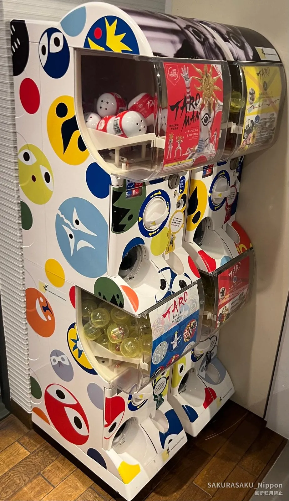 Gachapon capsule machines covered in Okamoto's face motifs, holding TAROMAN figures and badges