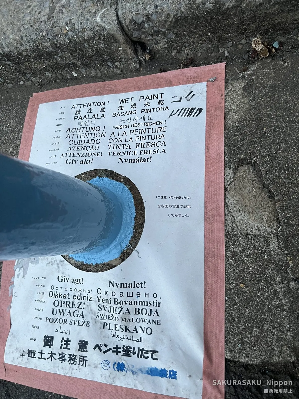 A home-made printed sheet taped to the road, listing "wet paint" in eighteen languages around a hole cut for the post