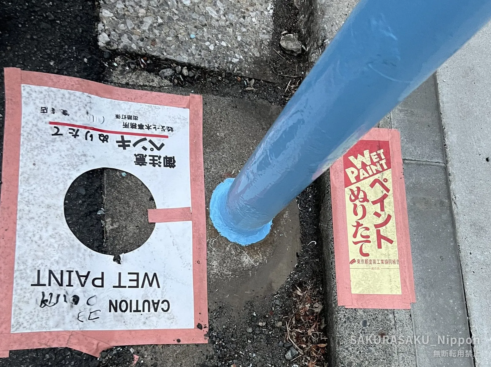 A freshly painted blue guardrail post, with a printed WET PAINT notice taped to the road beside it and a red-and-gold union tag on the kerb