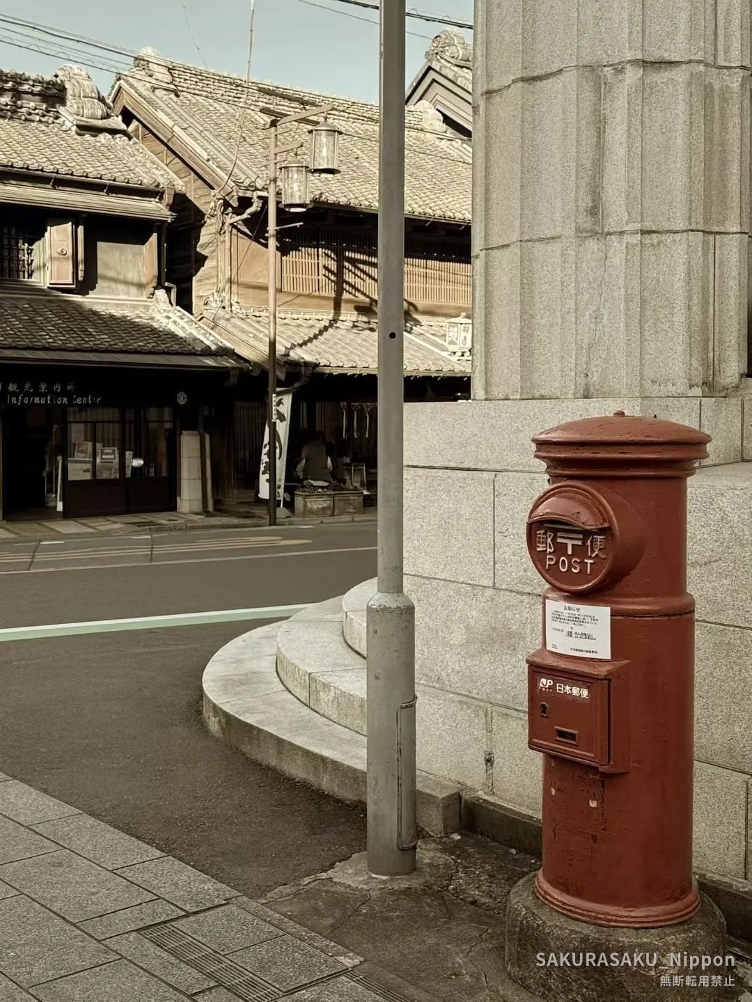 An old-fashioned red cylindrical postbox on a street corner in Kawagoe, with kura buildings and the tourist information centre behind