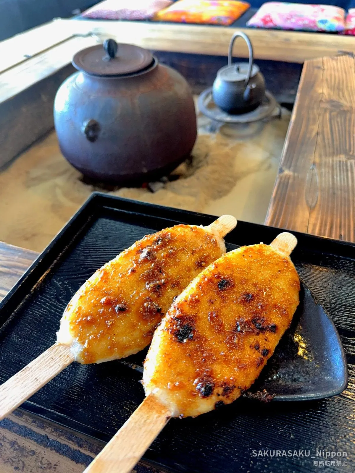 Two skewers of gohei-mochi, glazed and grilled, on a black tray beside a sunken hearth