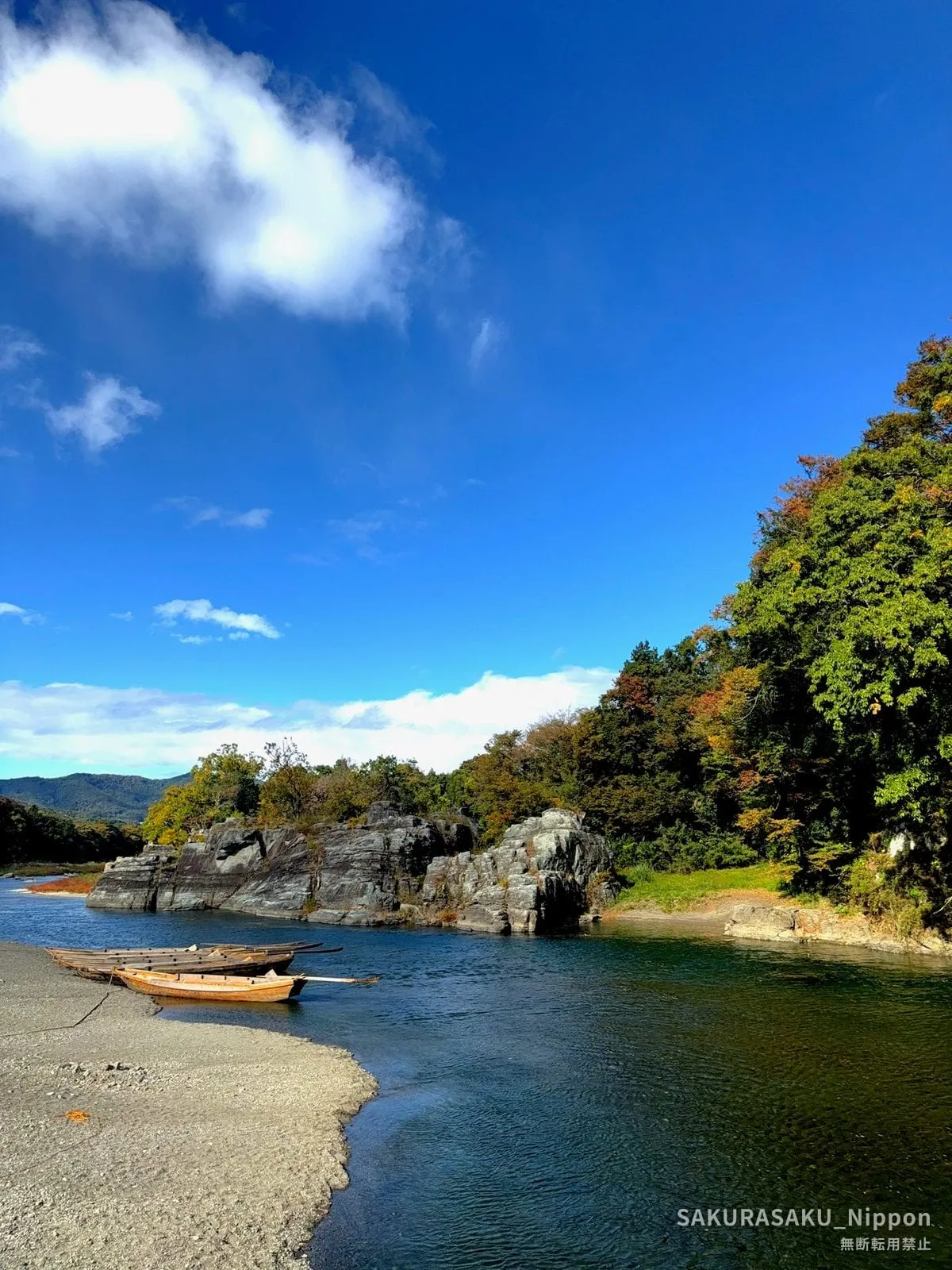Wooden line-kudari boats drawn up on the shingle beside the Arakawa, with wooded cliffs and autumn colour behind