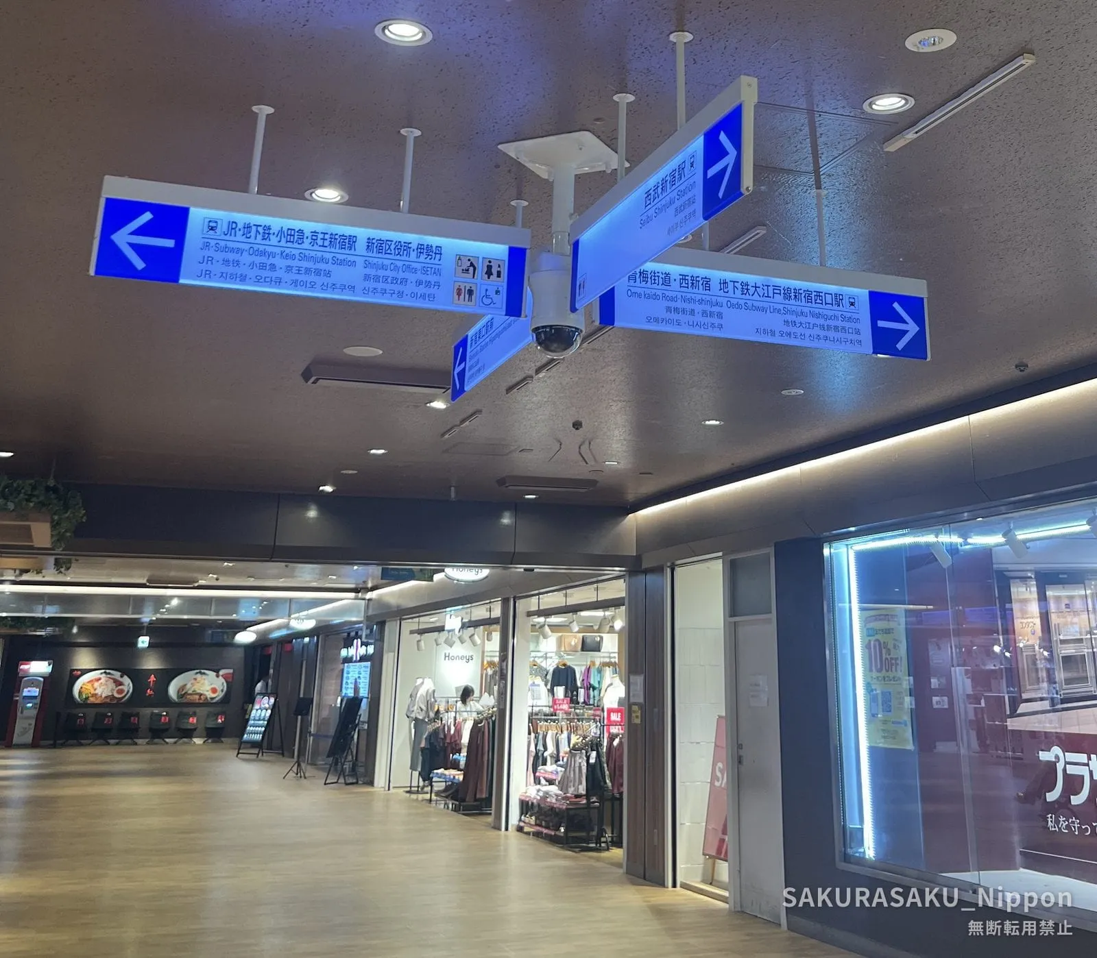 An overhead blue direction sign in four languages, pointing to Seibu Shinjuku Station one way and Ome Kaido, Nishi-Shinjuku and the Oedo Line the other